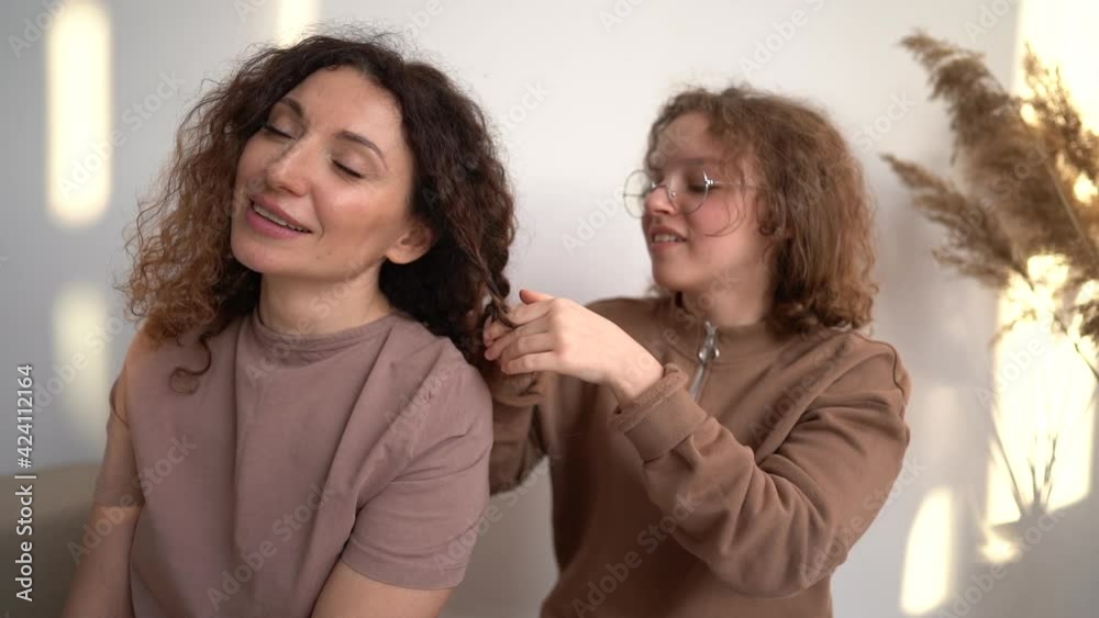 Portrait of mother and daughter at home. Teenage girl braids her lovely mature mother's hair Portrait of mother and daughter at home. Teenage girl braids her lovely mature mother's hair