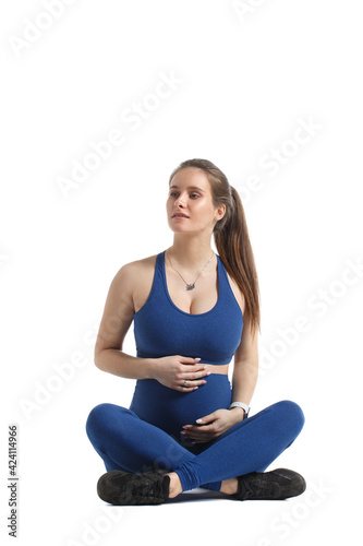 Cute pregnant woman in blue sportswear sits isolated on a white background.