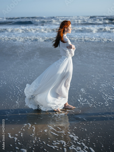 A woman in a white dress walks on the wet sand on the shore of the ocean in full growth