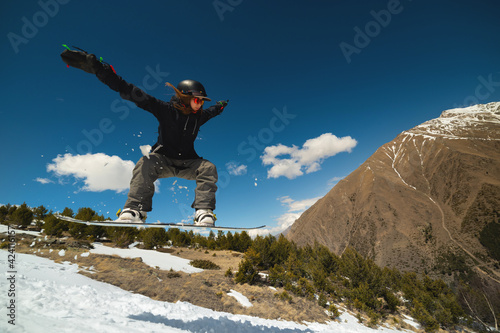 Snowboarder woman jumping from a kicker springboard from the snow on a sunny day in the mountains in a homemade snowboard park