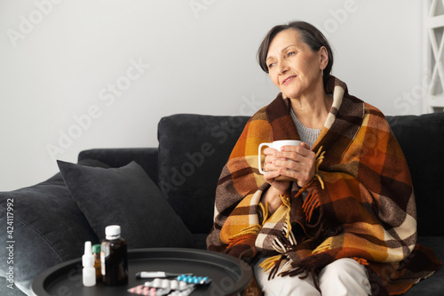 Smiling senior woman feels relieved, recovering after viral disease. An elderly female covered blanket sits on the couch, drinks hot and feels better. Healthcare concept