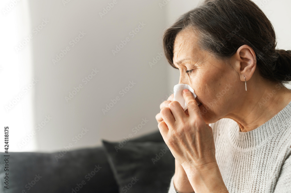 Sick senior woman sneezing, using a paper napkin for cleaning nose, has runny nose and cough, an older female got a viral infection, side view