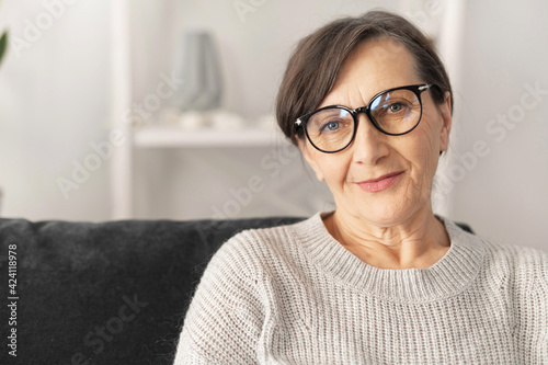 Charming middle aged woman wearing stylish eyeglasses looks at the camera with a calm friendly smile, close-up portrait of modern retire female indoor