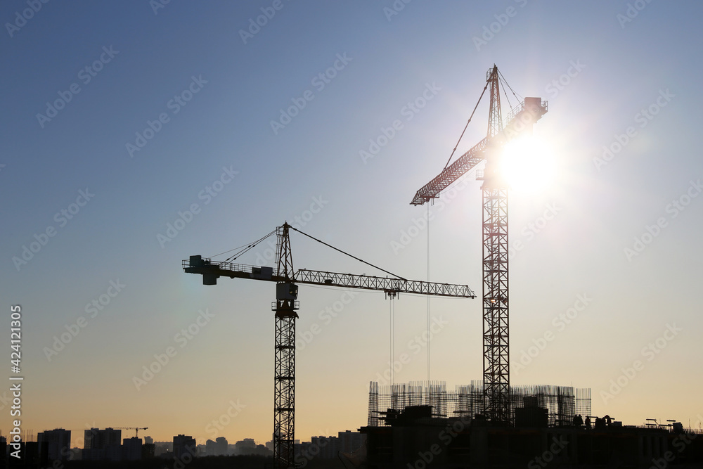 Silhouettes of tower cranes above the unfinished residential buildings against sunshine. Housing construction, apartment block in city