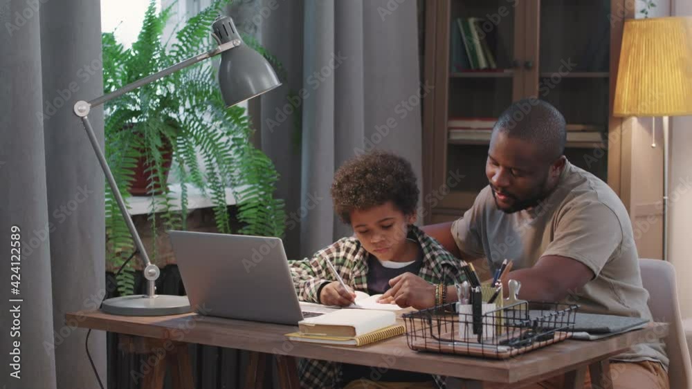 Panning medium shot of afro-american man helping his cute little son with school homework sitting together at desk in living room Panning medium shot of afro-american man helping his cute little son with school homework sitting together at desk in living room