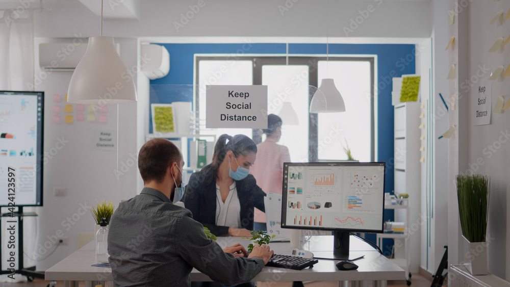 Freelancer with face mask working at financial graphics on pc sitting in business office. Coworkers talking in background about marketing mantain social distancing to prevent infection with covid19