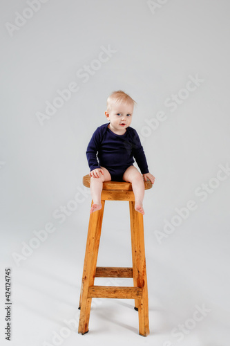 A child sitting on a wooden high chair, isolated against a gray background, a charming baby in a blue bodysuit, looks focused, waiting for his mother to play with him. The concept of childhood.