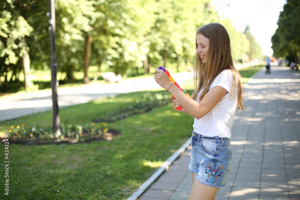 Naklejka premium Girl with a water pistol. Summer fun in the park with a children's water gun. Blonde with long hair in denim shorts and a white T-shirt. Play with water, shoot with water