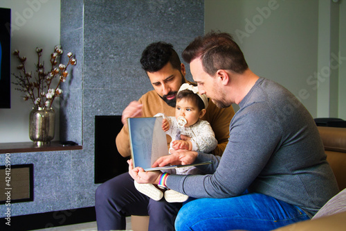 gay couple and their daughter sitting on a sofa reading a story to their little girl.