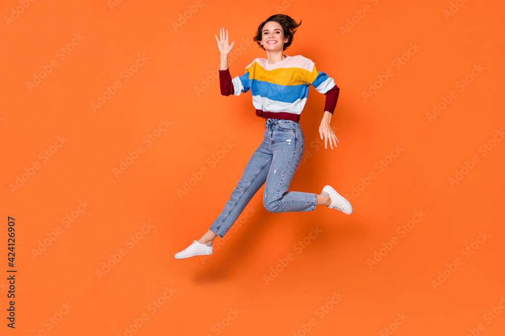 Photo of funky adorable young lady dressed striped pullover jumping waving arm isolated orange color background