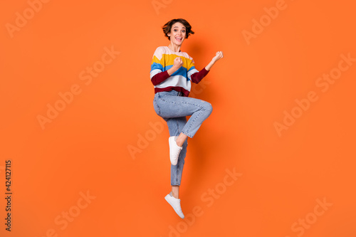 Photo of lucky shiny young lady dressed striped pullover jumping high rising fists isolated orange color background