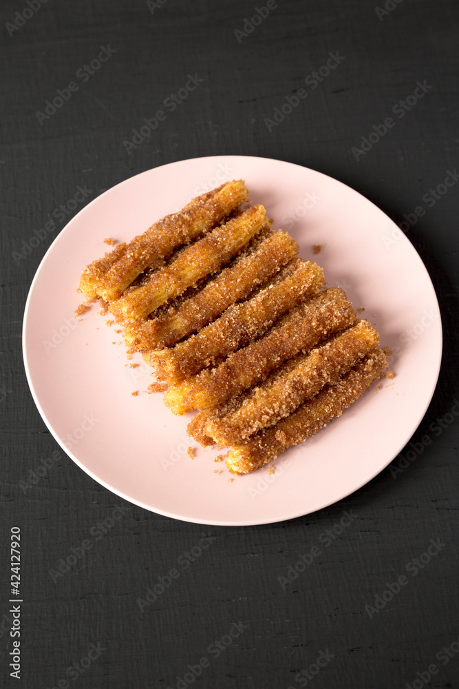Home-baked Churro Bites on a pink plate on a black surface, side view. Home-baked Churro Bites on a pink plate on a black surface, side view.
