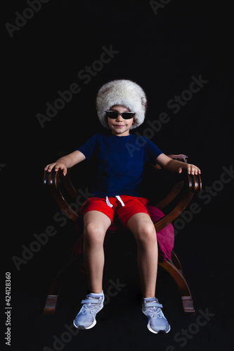 portrait of a child wearing a russian fur hat sitting on a wooden chair on black background