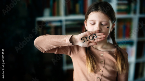 Stop - text on the palm of the hand and gesture. Heart on the cheek. Child girl is closed by hand with the inscription stop. Dark photo. Violence concept, stop violence and silence.