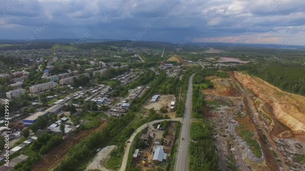 Aerial view of small Provincial industrial city. On the outskirts of the city there are low houses, garages and quarries. Cars drive along the road. Summer evening, in the sky doe clouds and blue clou