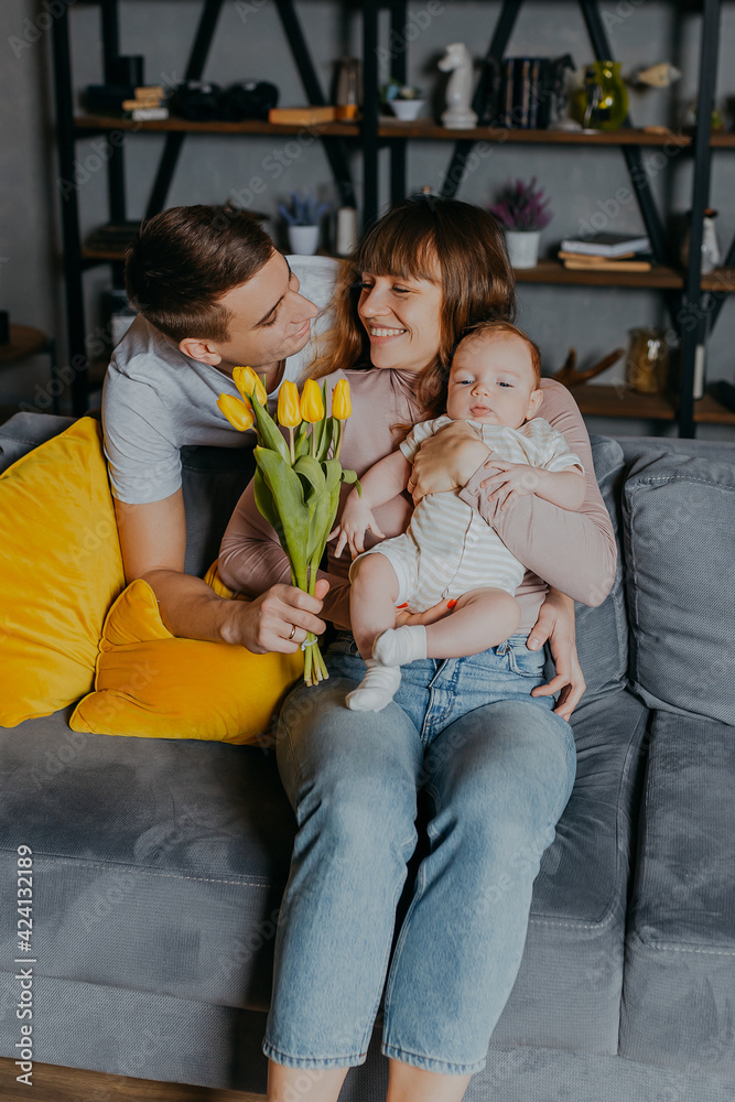 Man gives flowers to woman with little kid in arms. Mother's Day congratulations. Happy family with flowers on couch. Bunch of yellow tulips.