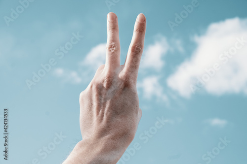 The victory sign with the fingers of the hand on the background of the blue sky. Two fingers are raised to the top against the sky.