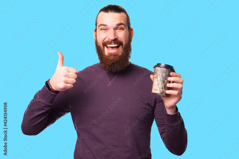 Young bearded man is smiling at the camera and showing a thumb up is holding a cup of hot drink Young bearded man is smiling at the camera and showing a thumb up is holding a cup of hot drink