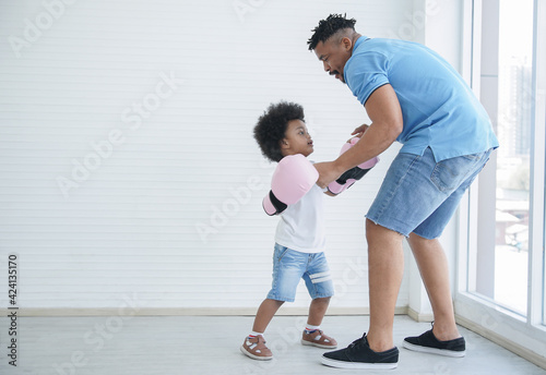 Dark skinned African father and little kid son playing with boxing gloves. Young dad with beard and child boy working out and fun together on white background at home near window  © Pruksachat
