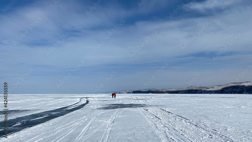 Two tourists walk on the ice of frozen Lake Baikal. Blue transparent ice with deep cracks, covered with snow.