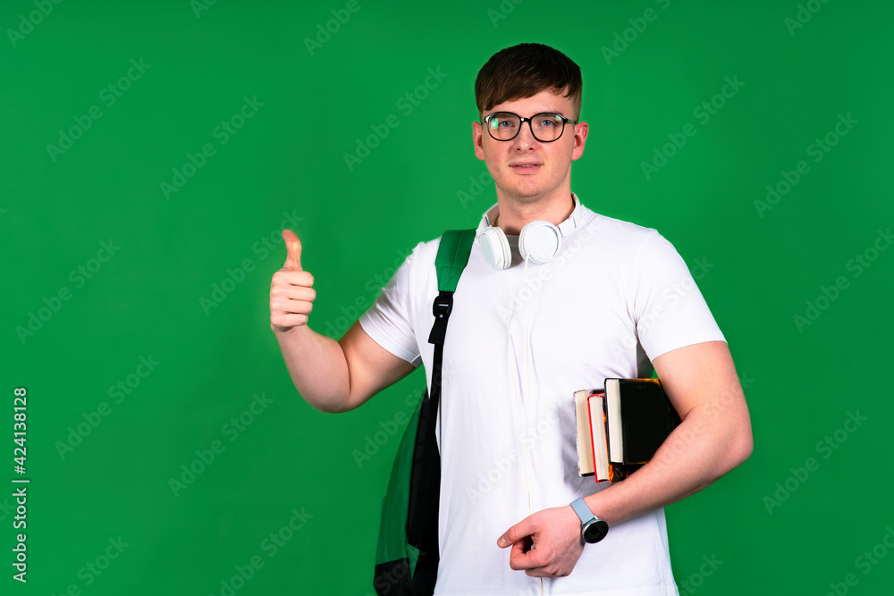 Handsome male student smiling and showing thumb up gesture, cool sign like. Happy guy in shirt outdoors, copy space. On green isolated background. Man holds textbooks and books in his hands.