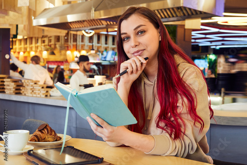 Hipster woman with red-dyed hair works remotely in a cafe, holding a notebook in her hands.