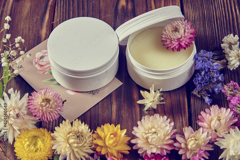 Handmade cream on a dark wooden surface surrounded by bright dried flowers. Handmade cream on a dark wooden surface surrounded by bright dried flowers.