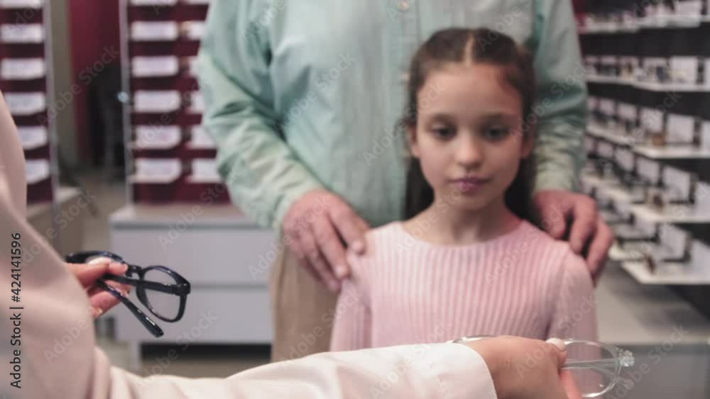 Medium closeup of little girl choosing pair of eyeglasses standing in optical store with grandfather Medium closeup of little girl choosing pair of eyeglasses standing in optical store with grandfather