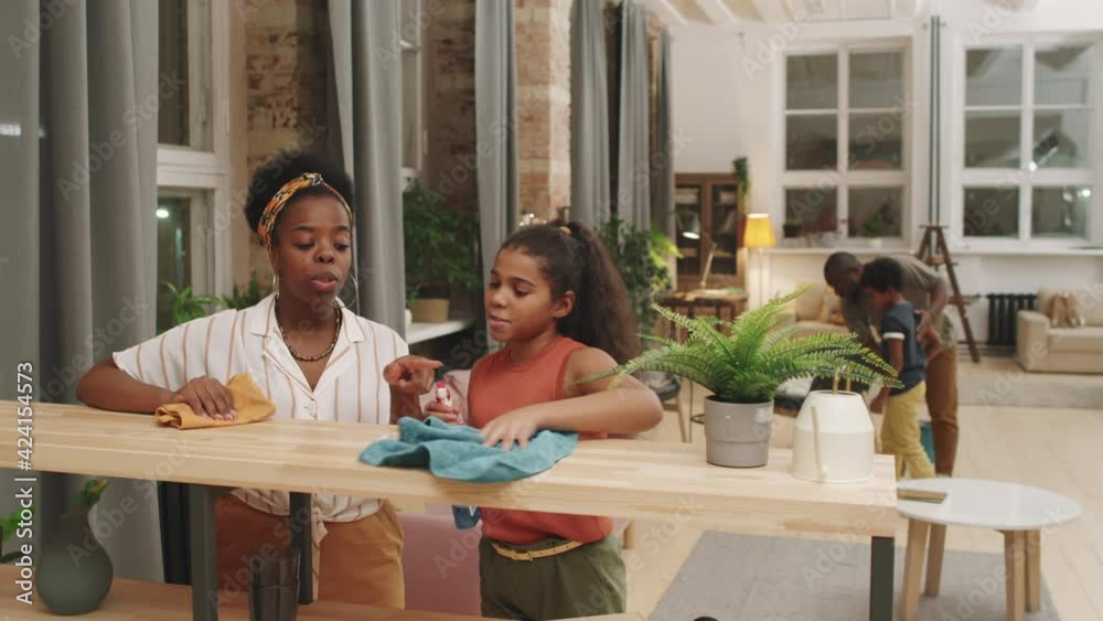 Medium shot of happy afro-american family of four chatting while doing housework in living room together. Girls dusting while boys cleaning floor in background Medium shot of happy afro-american family of four chatting while doing housework in living room together. Girls dusting while boys cleaning floor in background