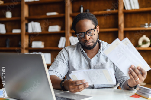 Serious black man with black square glasses, concentrated reading from whom letters, received a bad news, male manager employee unpacking banking notification, law order or paper document at workplace