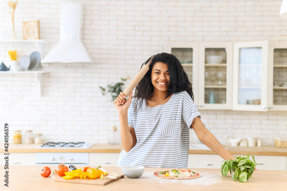 Young cheerful African American girl standing next to kitchen table and holding a rolling pin, concept of a woman in an everyday household routine, making cooking pizza, preparing dough and veggies