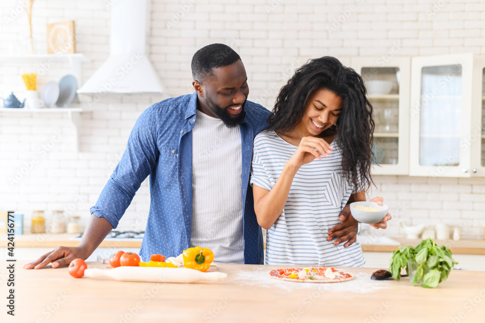 Overjoyed mixed-race couple boyfriend and girlfriend in love standing next to kitchen counter, hugging, spending leisure time weekend together at home, cooking, girl adding spices, seasoning pizza