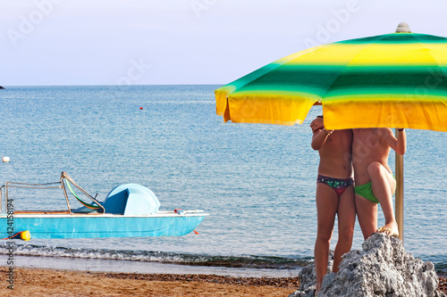 Two kids playing on a rock under the beach umbrella on the seashore.
