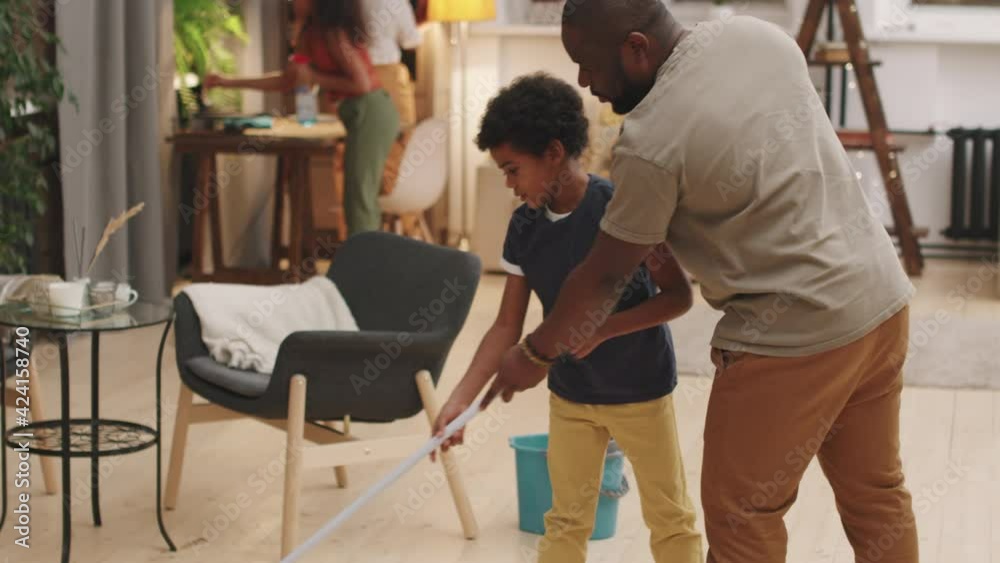 Tilting-up shot of cheerful afro-american family of four doing housework in their apartment together. Father and cute little son mopping floor while mom and daughter dusting in background