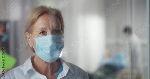 Female mature doctor in face mask in hospital standing near window