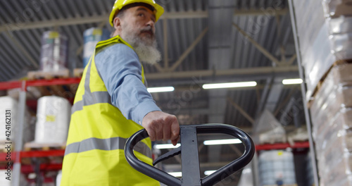Senior worker in warehouse using hand pallet stacker to transport goods.