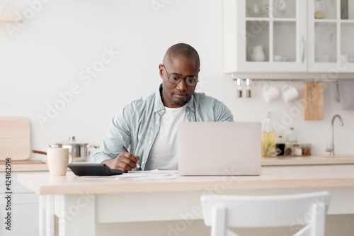 Budget planning concept. Pensive african american man calculating taxes and using laptop computer, sitting in kitchen