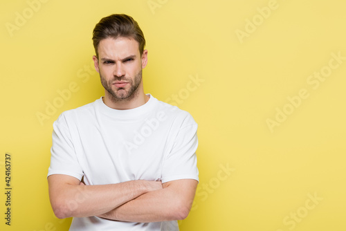 angry man looking at camera while standing with crossed arms on yellow