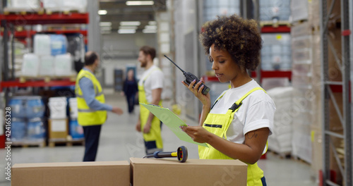 African female warehouse manager holding clipboard and using walkie-talkie while checking inventory. photo