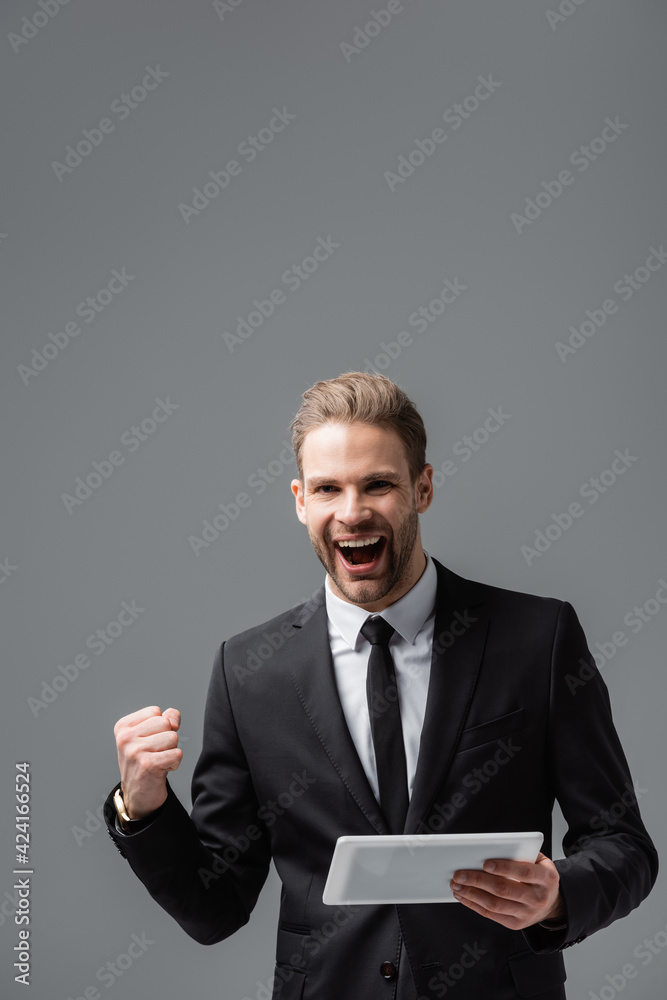 excited businessman showing success gesture while holding digital tablet isolated on grey excited businessman showing success gesture while holding digital tablet isolated on grey