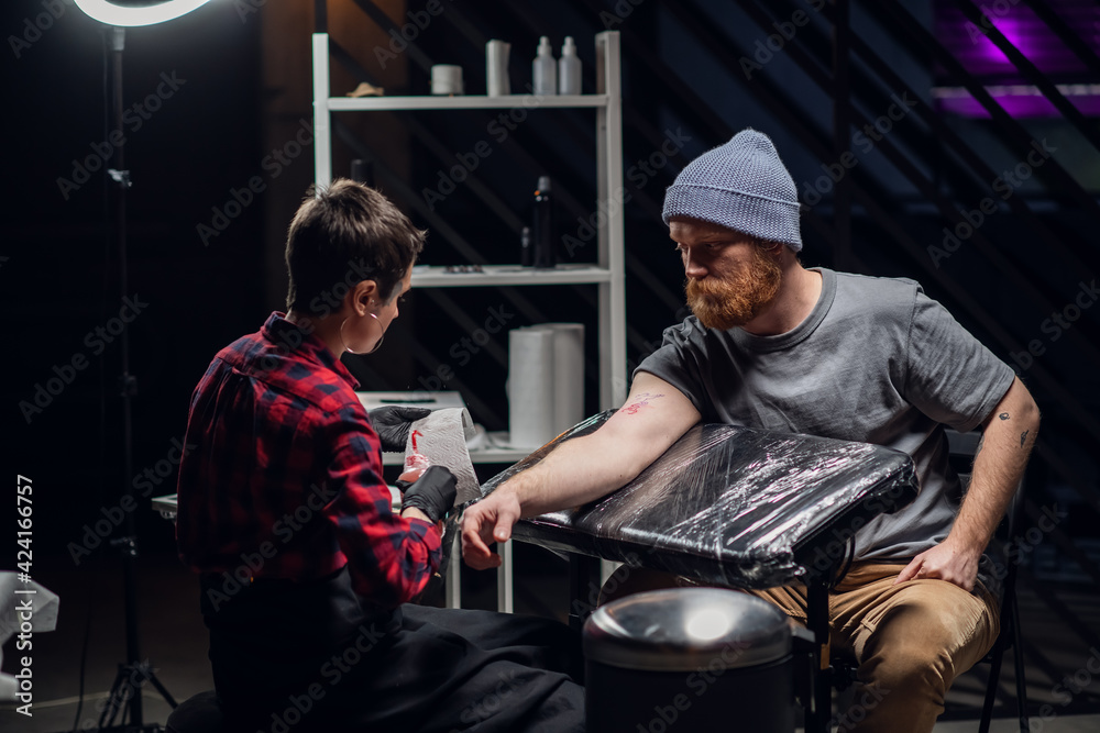 In a modern tattoo parlor, a girl with piercings and tattoos on her body makes a tattoo on the bicep of a young hipster's hand.