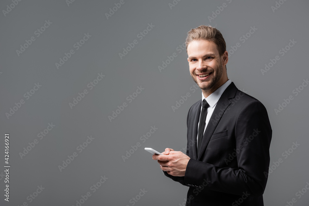 positive businessman smiling at camera while chatting on smartphone isolated on grey