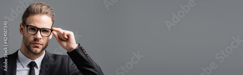 young, serious businessman adjusting eyeglasses while looking at camera isolated on grey, banner