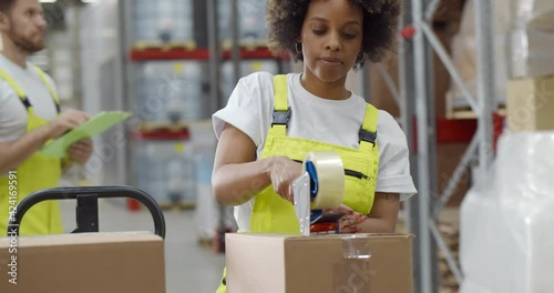 African female worker packing cardboard box with tape gun dispenser in warehouse photo
