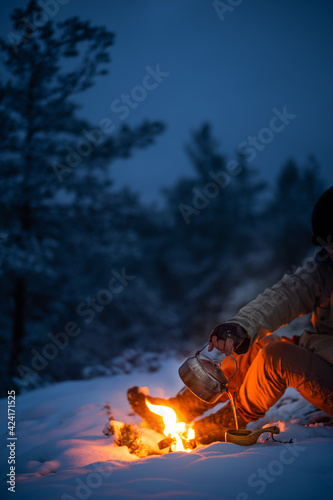 Man sitting in a snow-clad forest pouring coffee made on woodfire into a wooden cup.