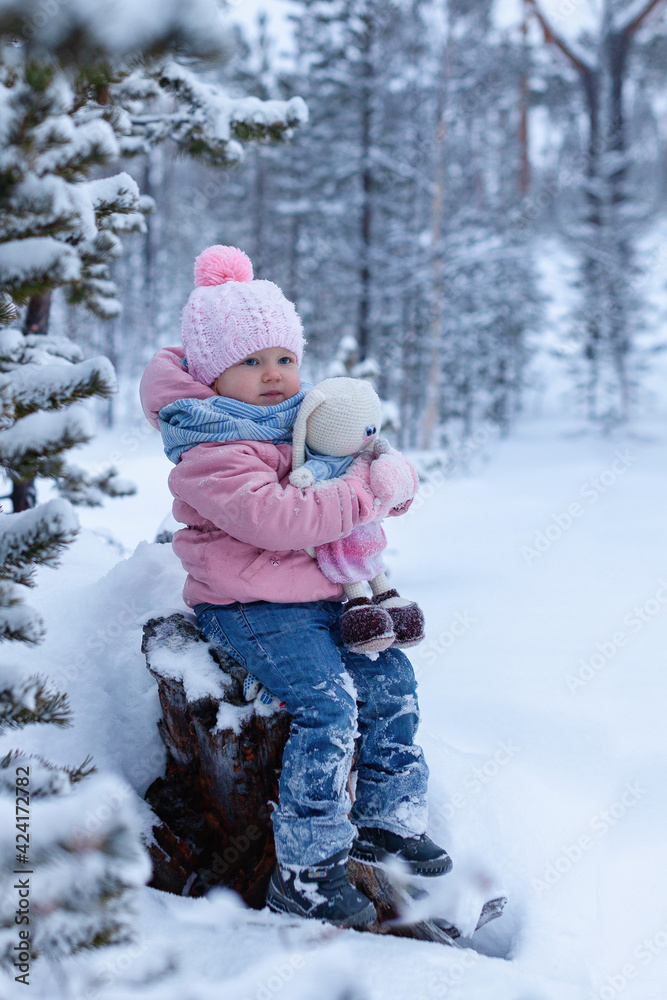 Toddler little girl sits on a stump in snowsuit on a winter day. hugs a knitted toy