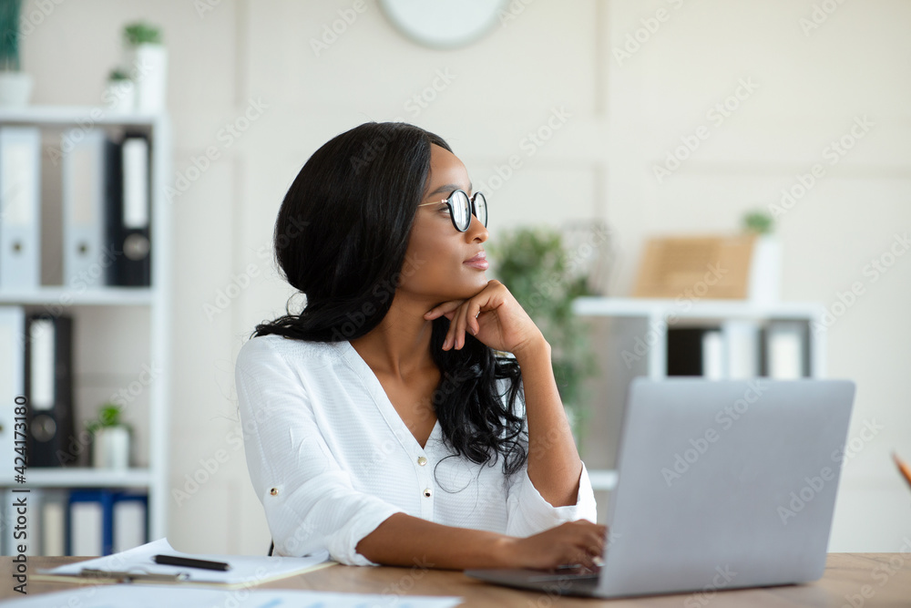Millennial black entrepreneur daydreaming in front of laptop at contemporary office Millennial black entrepreneur daydreaming in front of laptop at contemporary office