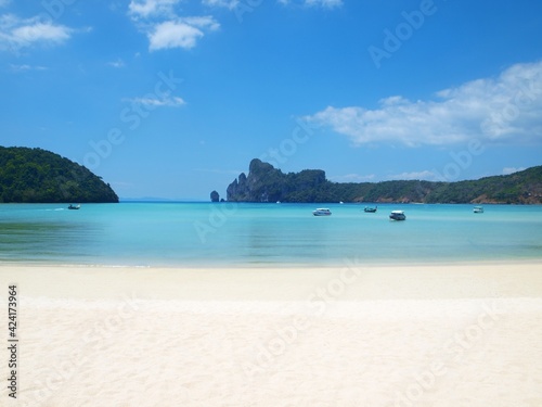 Small deserted beach in a sea bay. White clean sand, transparent bright turquoise water, rocks covered with green forest on the horizon. Boats on the calm water. Thailand, tropical resort. Beautiful