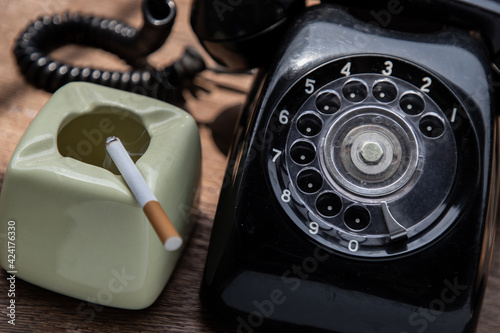 Cigarette that were burning placed on greyish green ashtray and Old black retro rotary telephone on old wooden rustic table. Copy space, Selective focus.