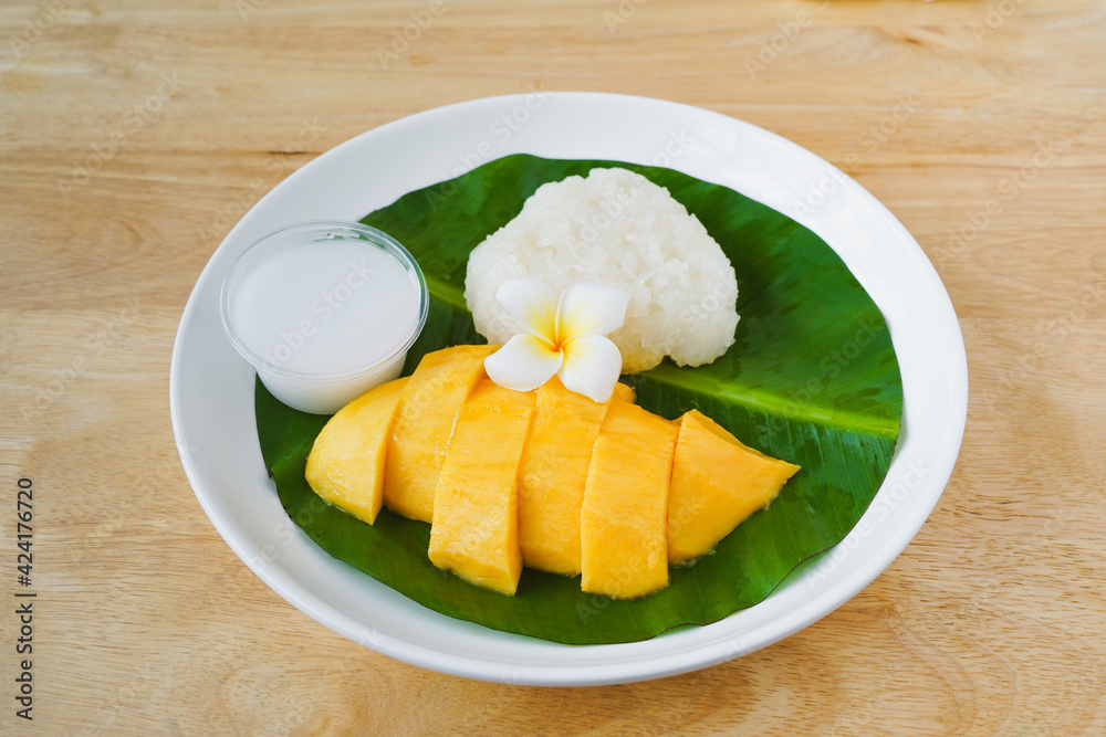 sweet ripe mango , sticky rice and coconut milk in small bowl on green banana leaf and white plate with blurred wood desk including clipping path around plate easy to remove background sweet ripe mango , sticky rice and coconut milk in small bowl on green banana leaf and white plate with blurred wood desk including clipping path around plate easy to remove background